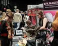 A bustling convention scene features attendees engaged in various activities, with a focus on horror themes. In the foreground, a woman dressed in a horror-themed outfit examines items on a table, while another attendee, wearing a brown hat, stands nearby. The table displays an array of props, including a large, intricate creature model, and promotional materials for horror-related events and products. The background is filled with other convention-goers and vendor booths, enhancing the immersive atmosphere of the event.