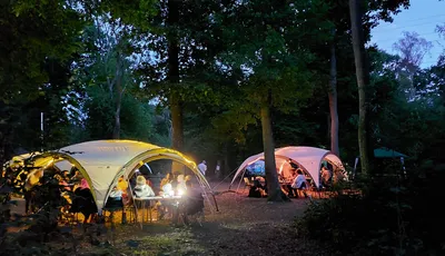 Two large, white canopies are set up in a wooded area during dusk, illuminated by warm lights. Under one canopy, a group of people sits around tables, engaged in conversation and activities, while another canopy hosts a similar gathering. The surrounding trees create a serene atmosphere, enhancing the friendly and inclusive vibe of the event.