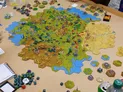 A detailed tabletop gaming scene features a large hexagonal map covered in various terrain types, including green fields, golden hills, and blue water. Numerous miniature figures are positioned across the map, with flags indicating different territories, and several dice are scattered nearby. In the foreground, a notepad and a collection of dice in various colors are visible, suggesting active gameplay.