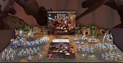 A large, detailed box titled "Grimdark Future: Broken Truth" is prominently displayed at the center, surrounded by various miniatures and game components. To the left, a collection of colorful plastic miniatures representing the Saurian Starhost is arranged, while to the right, the Robot Legions miniatures are showcased. Additional items include a quickstart guide, measuring tools, dice, and full-color terrain pieces, all set against a backdrop of a gaming mat and themed scenery.