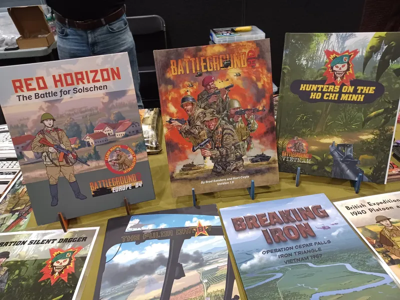 A display of wargaming rulebooks and supplements is featured on a table, showcasing various titles. Prominent among them is "Battleground HD," which depicts soldiers in action on its cover, alongside "Hunters on the Ho Chi Minh," featuring a jungle scene. Other titles include "Breaking Iron," "The Fulda Gap," and "British Expeditionary 1940 Platoon," each with distinct artwork reflecting military themes.