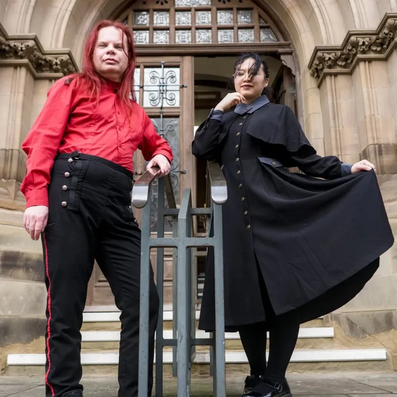 Two individuals pose in front of a grand, gothic-style building. One person, wearing a bright red shirt and black trousers with red stripes, stands confidently with arms crossed. The other person, dressed in a long black coat with a high collar and a blue shirt underneath, holds the hem of their coat, creating a dramatic silhouette. The ornate entrance of the building, featuring large wooden doors and intricate stonework, serves as a striking backdrop.