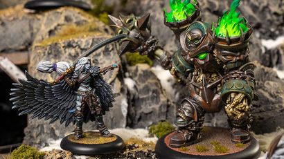 A detailed miniature scene features a character with large, intricately designed black wings, dressed in a mix of armor and clothing, wielding a weapon and casting a spell. Opposite this character stands a massive, armored figure with glowing green accents and a spiked weapon, set against a rocky terrain with patches of moss. The contrasting sizes and designs of the two figures highlight the dynamic nature of the scene.