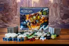 A G.I. JOE Heroscape: Rumble at the Rift Battle Box is prominently displayed in the center, featuring vibrant artwork of characters in action. Surrounding the box are various game components, including eight miniatures representing characters like Duke and COBRA Commander, modular hex terrain pieces, COBRA fortifications, dice, and markers. The background features a blurred, crumbling environment, enhancing the game's adventurous theme.