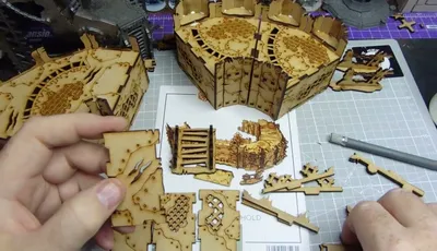 A hand holds a small, intricately cut piece of laser-cut wood, featuring detailed patterns and textures. Surrounding the hand are larger assembled sections of similar wooden terrain pieces, showcasing various designs, including grids and panels. In the background, a printed layout with additional terrain components is visible, indicating a crafting or gaming setup.