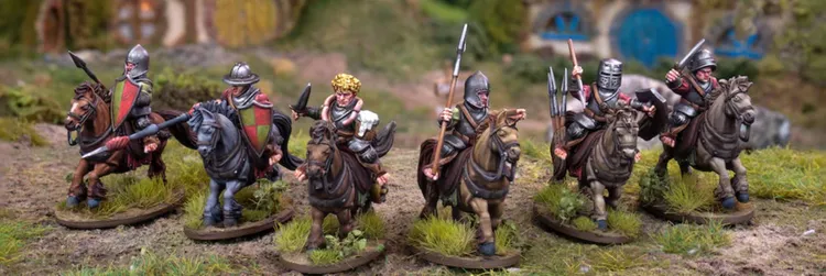 Four mounted halfling warriors are depicted, each riding stout ponies. The first rider has curly hair and holds a mug, while the second is clad in armor and wields a spear. The third rider, also armored, carries a spear and has a shield, and the fourth rider is similarly equipped. The scene is set against a lush, grassy background, with hints of a rustic building in the distance.
