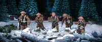 Three British and Canadian winter infantry figures are depicted in a snowy landscape, surrounded by patches of grass and rocks. Each figure is dressed in warm winter gear, with various poses: one holds a rifle, another has a sidearm, and the third is equipped with a machine gun. The background features dark green trees, enhancing the wintery atmosphere of the scene.