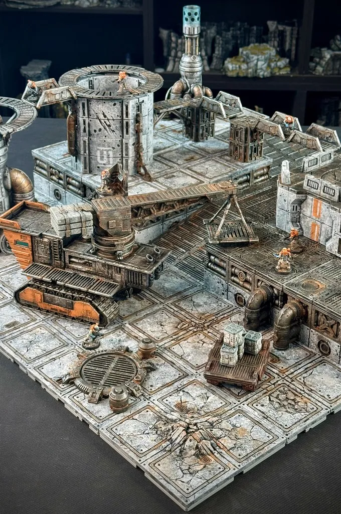 A detailed modular sci-fi terrain setup features a large, industrial-looking vehicle with tracks and a raised platform, surrounded by various structures and pipes. In the scene, two miniature figures are positioned on the terrain, one near a stack of crates and the other on a walkway. The ground is textured with cracked tiles and a circular grate, enhancing the gritty, futuristic atmosphere of the setting.