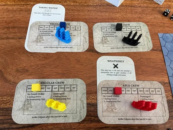 Four ship status cards are displayed on a wooden surface, each featuring a different color ship token: blue, yellow, black, and red. The cards provide information about the crew's condition, including statuses like 