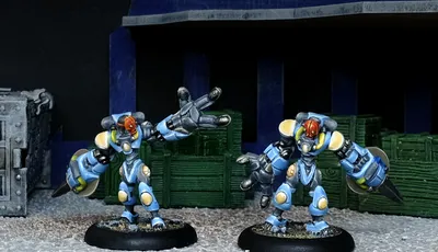 Two Assault Androsynth miniatures are prominently displayed in the foreground, designed for hand-to-hand combat. They feature a blue and yellow color scheme, with one holding a Vibro Scythe and the other a Cesti, showcasing their imposing, armored forms. The background includes dark crates and a textured surface, enhancing the urban warfare theme.