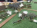 A detailed tabletop wargaming scene features miniature soldiers arranged on a green felt battlefield, with two small stone houses in the background. Various units, including infantry and artillery, are positioned strategically, with some figures holding rifles and others mounted on horses. Puffs of cotton represent smoke effects, adding to the dynamic atmosphere of the game.