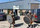 Two individuals dressed in historical warrior attire stand outside a building, each holding a round shield and a spear. They wear chainmail and tunics, with one having a long beard and the other a shorter beard. In the background, a sign for "Herefordshire Mind" is visible, along with a few parked cars and a large van.