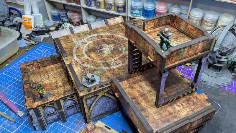 A detailed tabletop gaming scene features a multi-level terrain piece with a weathered, industrial design. Two miniature figures are positioned on the upper levels: one in a yellow and black outfit and another in a more rugged appearance, both poised on the edges of the structure. The background includes various hobby supplies, such as paint bottles and tools, indicating an active workspace.