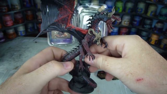 A detailed dragon miniature is being held in a hand, showcasing its intricate features, including prominent scales, fierce facial expression, and large wings with a red underside. The dragon is painted in dark hues with highlights that accentuate its muscular form and menacing appearance. In the background, various paint jars are visible, indicating a workspace for model painting.