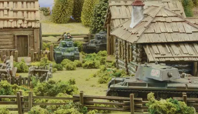 A detailed tabletop scene features several miniature tanks amidst a rural setting, including a wooden cabin and lush greenery. In the foreground, a tank with the number "600" is positioned near a wooden fence, while another tank is seen further back with a crew member visible. The background includes trees and additional structures, creating a vibrant battlefield atmosphere.
