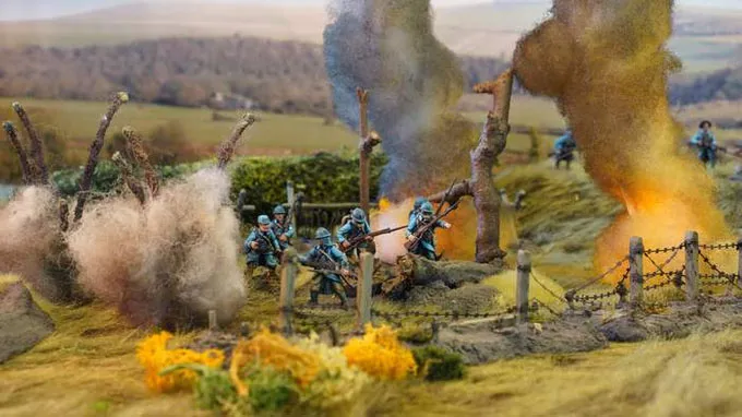A detailed diorama depicts a World War I battlefield scene with soldiers in blue uniforms engaged in combat amidst smoke and explosions. In the background, a landscape of rolling hills is visible, with barbed wire and trees partially obscured by billowing smoke. The foreground features vibrant yellow foliage, enhancing the dramatic atmosphere of the scene.