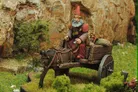A miniature figure of a gnome with a long beard and a red pointed hat sits atop a wooden cart pulled by a dark brown donkey. The cart is laden with various items, including sacks and a small barrel, and is set against a backdrop of rocky terrain and green foliage. The scene captures a whimsical, fantasy-inspired atmosphere, perfect for a tabletop RPG setting.