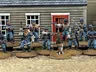 A group of ten detailed 28mm scale miniatures representing dismounted US Cavalry and Buffalo Soldiers stands in front of a wooden building with a red door. The miniatures are posed with various weapons, including rifles and a bugle, showcasing a mix of uniforms and accessories, such as scarves and hats. A small beagle and a cactus are also present among the figures, all mounted on 25mm round bases with a sandy terrain effect.