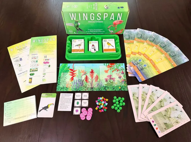 The Wingspan: Americas Expansion box is prominently displayed, featuring vibrant artwork of a hummingbird and a flamingo. Surrounding the box are various game components, including bird cards, a green tray for organizing pieces, and colorful goal cards. A beautifully illustrated game board depicting tropical flora is also visible, along with tokens and player boards, all set against a dark wooden table.