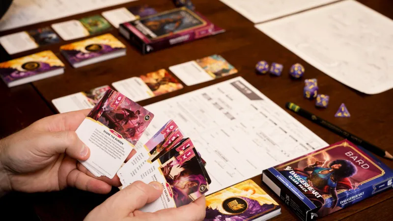 A tabletop gaming scene features a wooden table with various gaming materials. Prominently displayed is the "Daggerheart Class Pack" for the Bard, showcasing vibrant artwork on its cover. Nearby, a player holds a stack of character cards, while several polyhedral dice and character sheets are scattered across the table, indicating an active game session.
