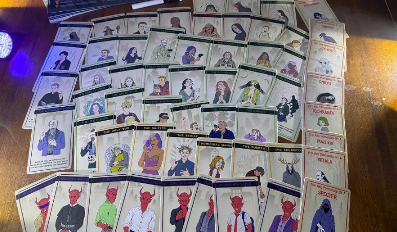 A collection of game cards from the social deduction game "Possess Me, Satan" is spread out on a wooden surface, showcasing a variety of characters. The cards feature illustrations of diverse individuals, including several with red-skinned, horned figures representing demonic characters, alongside others depicting humans in various poses and expressions. The cards are arranged in a somewhat chaotic manner, with some titles visible, hinting at the game's themes of possession and ghost hunting.