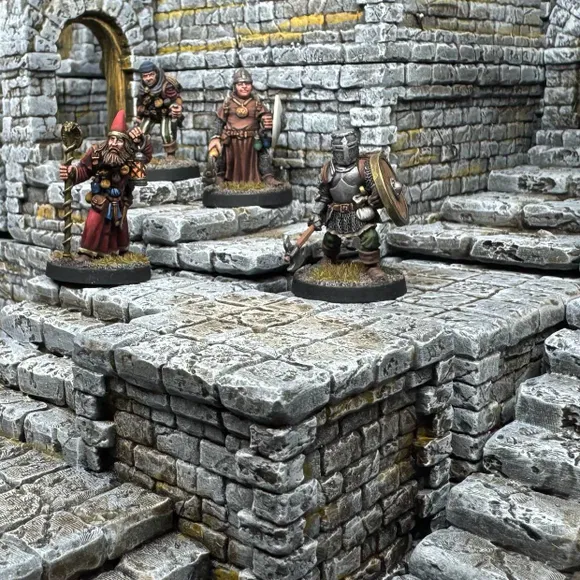 A detailed scene showcases a modular dungeon setup made from intricately sculpted stone blocks, featuring various levels and staircases. Four miniatures are positioned on the blocks: a bearded wizard with a staff, a robed figure holding a spear, and a knight in armor wielding a weapon, all engaged in exploration. The textured stone surfaces and the overall layout highlight the versatility of the dungeon blocks for tabletop gaming.