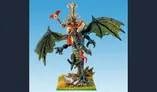 A detailed miniature depicts a liche king riding a zombie dragon. The liche king, adorned with skulls and a flowing red robe, holds a staff topped with a skull in one hand and a book in the other. The dragon features tattered wings and a menacing expression, with a long, serpentine body that includes skeletal elements. The base is grassy with rocky details, enhancing the undead theme of the scene. The background is a gradient of blue, emphasizing the figures in the foreground.
