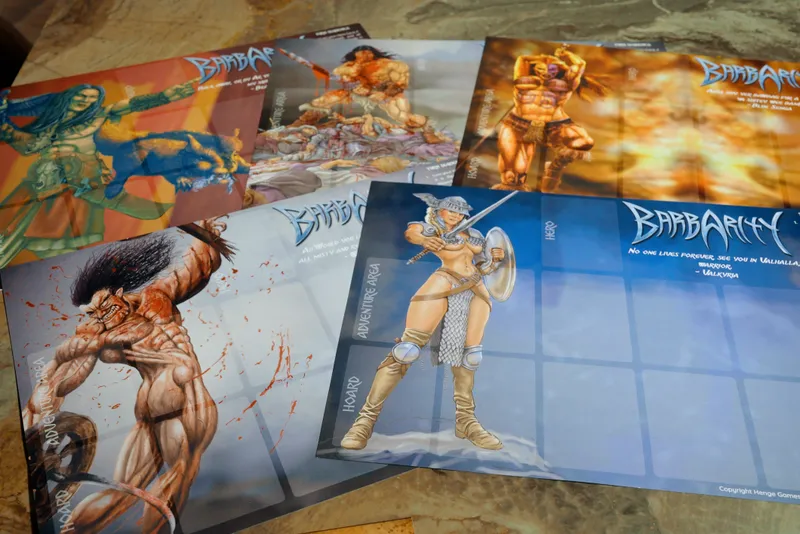 The image features several game components from the card game "Barbarity." Prominently displayed are character and gameplay boards, showcasing vibrant artwork that reflects a fantasy theme. One board depicts a muscular female warrior in minimal armor, wielding a sword and shield, while others illustrate various characters and scenes, including a bloodied background and a creature. The overall aesthetic channels classic fantasy illustration styles, with bold colors and dynamic poses, emphasizing the game's themes of combat and adventure. The boards are arranged on a textured surface, enhancing the visual appeal of the game components.