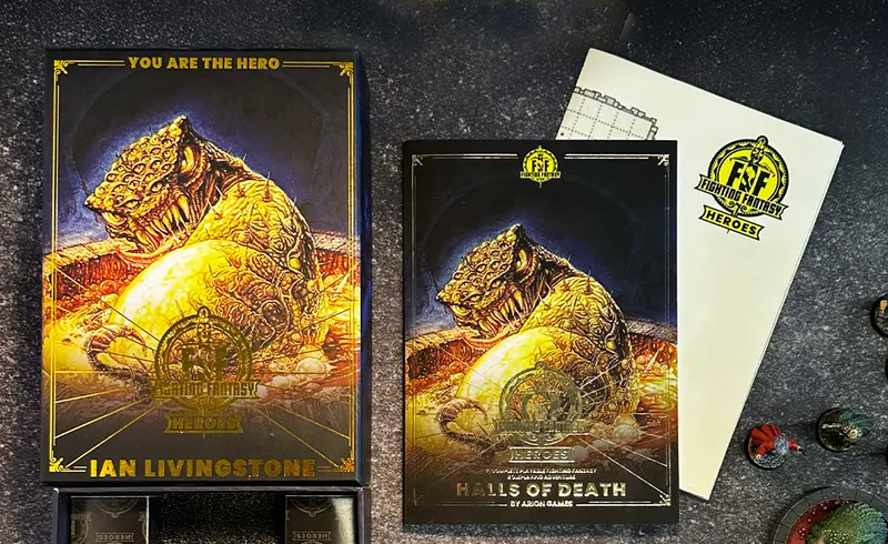 A beautifully designed box for "Fighting Fantasy Heroes" features intricate artwork, prominently displaying a creature with a textured, golden appearance. Next to it, an RPG adventure booklet titled "Halls of Death" showcases a similar creature on its cover, along with the Fighting Fantasy logo. Scattered around are various miniatures and a sheet with a dungeon map, all set against a dark, textured surface.