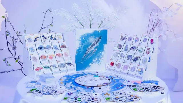 Boardgame components on a white table: Arctic wildlife cards showing fish, seals, whales, and birds; a circular game board with a blue Arctic map; hexagonal tiles with animal icons; and small circular tokens in blue, green, and purple. Central artwork depicts a blue whale.