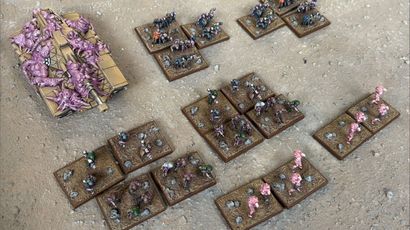 A collection of Zayken miniatures from Void Scar Miniatures is displayed on a textured surface. In the foreground, several bases feature hordes of zombified figures in various colors, including green, purple, and pink, representing the Neophyte Mobs. To the left, a tank is adorned with grotesque, bloated creatures, suggesting a fusion of machine and undead, while additional bases of zombies are scattered throughout the scene, emphasizing the overwhelming numbers of the Zayken forces.