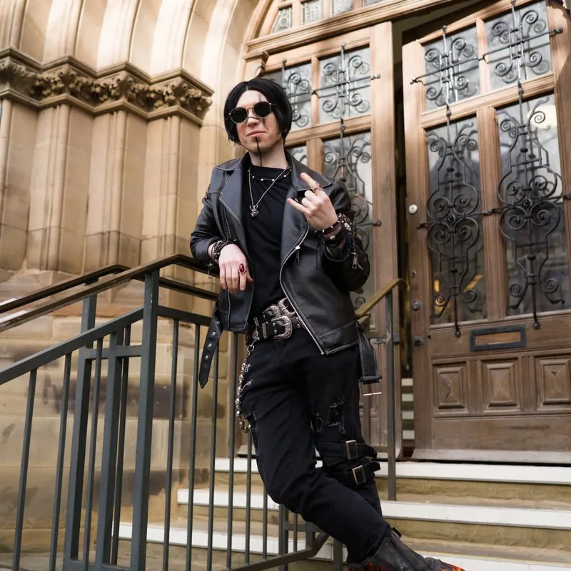 A person dressed in a black leather jacket and dark clothing stands confidently on a staircase, striking a rock-and-roll pose with one hand raised in a gesture. They wear sunglasses and have a stylish, short hairstyle, complemented by accessories like chains and rings. In the background, ornate wooden doors with decorative ironwork are visible, hinting at the gothic architecture of the venue.