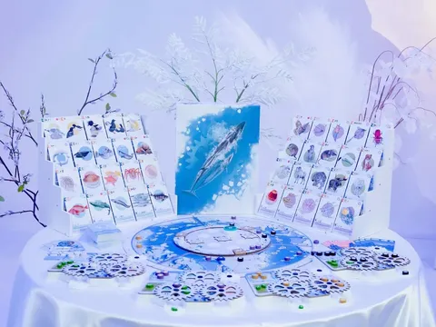 Boardgame components on a white table: Arctic wildlife cards showing fish, seals, whales, and birds; a circular game board with a blue Arctic map; hexagonal tiles with animal icons; and small circular tokens in blue, green, and purple. Central artwork depicts a blue whale.