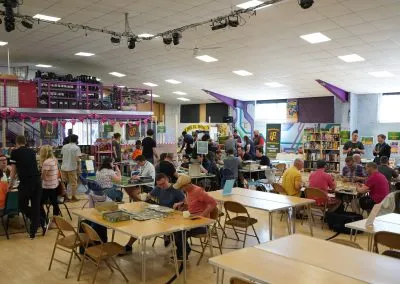 A spacious gaming hall filled with tables and chairs, where groups of people are engaged in various tabletop games. In the background, shelves are stocked with board games, and a vibrant display area features colorful banners and decorations. Attendees are seen socializing and playing games, creating a lively and inclusive atmosphere.