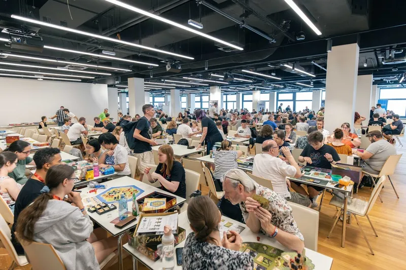 A bustling scene at Austria's largest games event, featuring a spacious hall filled with rows of tables where attendees are engaged in various board games. Many participants are seated, some focused on their games while others interact with fellow gamers, creating a lively atmosphere. The room is well-lit with large windows, and numerous game boxes are visible on the tables, showcasing a diverse selection of games available for play.