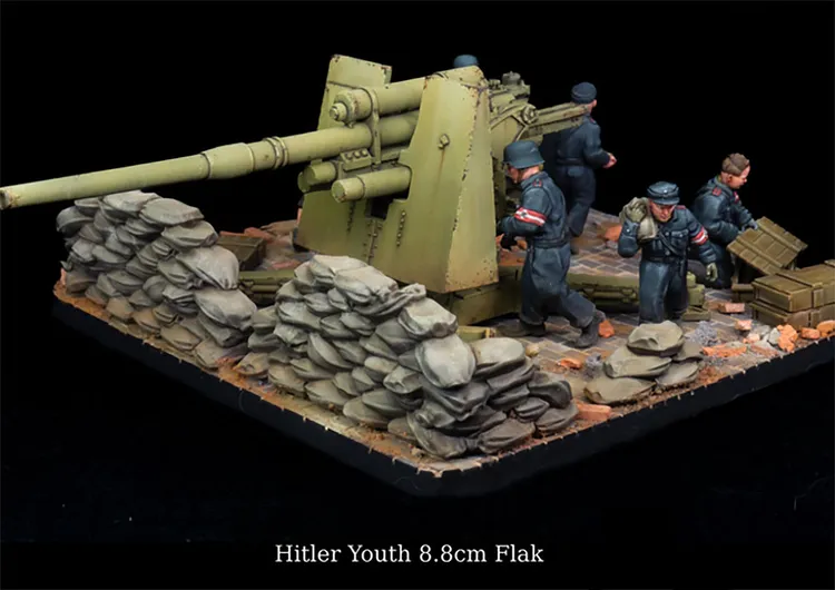 A detailed miniature scene features a 8.8cm Flak gun positioned behind a barricade of sandbags, with several figures representing Hitler Youth soldiers in various poses. The soldiers are dressed in dark uniforms with red armbands, engaged in their roles around the artillery piece. The base is textured with rubble and debris, enhancing the wartime atmosphere of the diorama.
