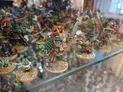 A display case filled with a variety of painted miniature figures, primarily featuring green-skinned goblins and orcs. The foreground showcases a larger orc wearing armor and holding a sword, alongside smaller goblins in playful poses. The background is populated with more miniatures, including various fantasy-themed characters and creatures, all set against a textured base that mimics a natural terrain.