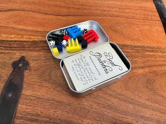 A small, silver tin box is open on a wooden surface, revealing the contents of the Battle Box - Age of Sail game. Inside, there are colorful plastic tokens in red, blue, yellow, and black, along with four dice. A stack of cards, labeled 