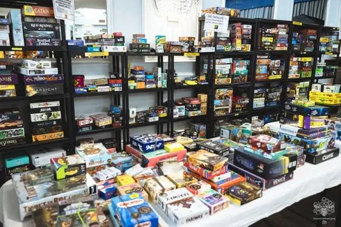 A vibrant display of board games fills a large table in the foreground, showcasing various game boxes with colorful artwork. Behind the table, several black shelves are lined with even more board games, creating a rich tapestry of titles and designs. The setting appears to be a bustling event space, likely part of the Manchester Board Games Festival, with a bright and inviting atmosphere.