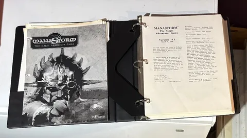 A three-ring binder containing the playtest manuscript for "ManaStorm: The Magic Adventure Game," featuring a black and white cover illustration of a character with a spiked helmet and a weapon. The title "MANASTORM The Magic Adventure Game" is prominently displayed at the top, along with the version number and date on the first page inside the binder. Additional pages include loose character sheets and player handouts, indicating the game's mechanics and spells.