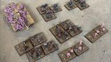A collection of Zayken miniatures from Void Scar Miniatures is displayed on a textured surface. In the foreground, several bases feature hordes of zombified figures in various colors, including green, purple, and pink, representing the Neophyte Mobs. To the left, a tank is adorned with grotesque, bloated creatures, suggesting a fusion of machine and undead, while additional bases of zombies are scattered throughout the scene, emphasizing the overwhelming numbers of the Zayken forces.