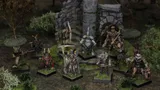 A group of intricately detailed fantasy miniatures stands in a lush, green setting near a stone ruin. The figures include various characters such as a skeletal leader holding a staff, a warrior with a bow, and a creature resembling a humanoid animal, all showcasing vibrant colors and classic 80s-style designs. Surrounding them are patches of grass and small bushes, enhancing the scene's immersive tabletop atmosphere.