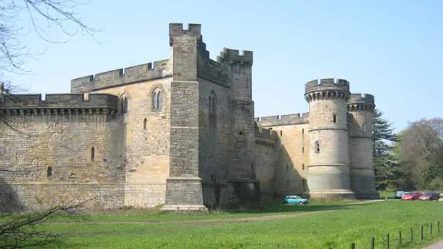A large stone castle with multiple towers and thick walls, set against a clear blue sky. The castle features a mix of architectural styles, with rounded towers and crenellated parapets. In the foreground, a grassy area is bordered by a low fence, and a few cars are parked nearby.