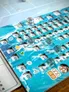 Board game with blue water-themed tiles. Grid of numbered hexagonal spaces, each with small icons: houses, boats, trash, and a circular island. Three translucent blue cubes on some spaces. Yellow and green pawns on the board.