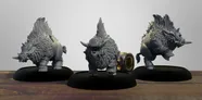 Three stylized miniature boars are displayed on black bases, showcasing their unique designs. Each boar features a thick, shaggy coat and prominent tusks, with one wearing a helmet and armor, suggesting a battle-ready appearance. The background is a simple, light color, allowing the details of the miniatures to stand out prominently.