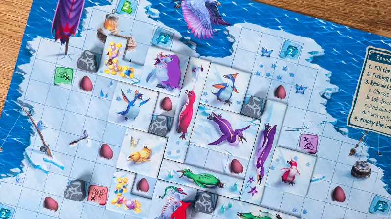 A colorful board game layout on a wooden surface. The game board features a grid of light blue icy spaces with some spaces containing gray rock tiles. Various illustrated penguin-shaped tiles in colors like purple, green, red, and blue are placed on the board. Pink egg-shaped tokens and gold coin tokens are scattered across the board. In the top left corner, there is a lantern token and a small wooden chest with gold coins spilling out. The top section of the board shows a large white and pink bird in flight over icy terrain and blue water. Some spaces on the board have icons with the number "2" and fish symbols. Small star-shaped tokens and additional rock tiles are also visible.
