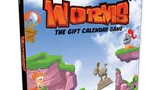 A colorful box for "Worms: The Gift Calendar Game" features a vibrant illustration of cartoonish landscapes with green hills and rocky cliffs. Prominent characters include a pink worm wearing a crown and a knight, both positioned on the cliffs, while a hot air balloon floats in the sky. The title is boldly displayed at the top, emphasizing the festive theme of the advent calendar.