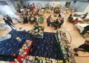 A spacious indoor venue filled with tables where numerous people are engaged in various tabletop games. In the foreground, several tables display a wide array of board games, with colorful boxes and game components visible. The background features groups of attendees playing games, chatting, and enjoying the event, with banners and signs indicating the convention's activities.