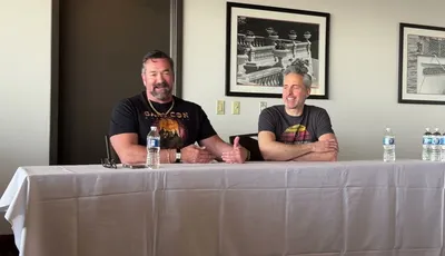 Two men are seated at a table during a panel discussion. On the left, a man with a beard, wearing a black shirt featuring a colorful graphic and a necklace, gestures with his hands while speaking. On the right, a man with short gray hair, dressed in a gray t-shirt with a retro design, smiles and listens attentively. Behind them, a framed black-and-white photograph of a decorative railing is visible on the wall. A bottle of water and a small device are placed on the table in front of them.
