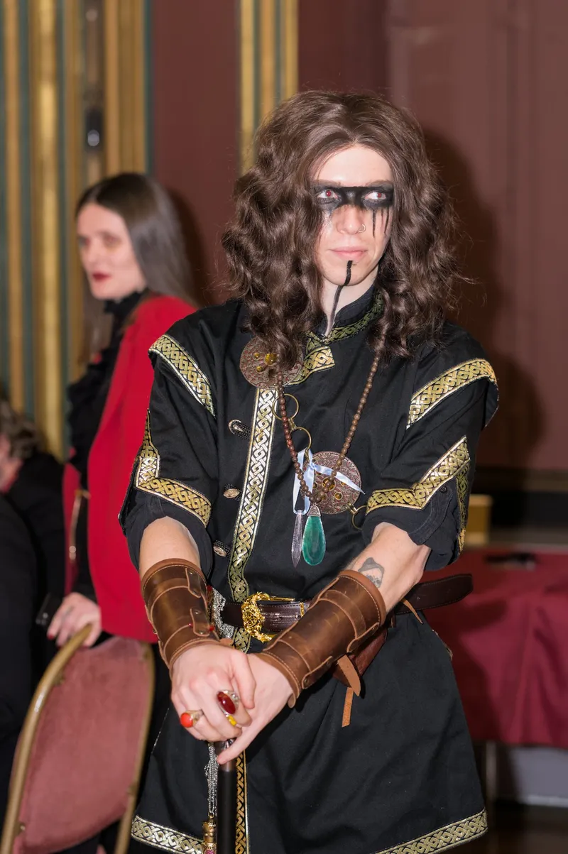 A person dressed in a dramatic vampire-themed costume stands in a gothic setting, featuring a long, dark robe adorned with intricate gold patterns. Their face is painted with striking black makeup around the eyes, and they have long, wavy hair. In the background, another individual in a red outfit is partially visible, contributing to the overall atmospheric vibe of the event.